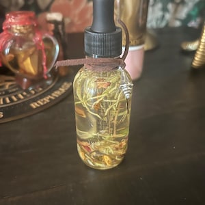 May include: A clear glass bottle with a black dropper top, filled with a yellow liquid and botanical elements. A brown cord is wrapped around the neck, and a silver charm hangs from it. The bottle is on a dark surface.