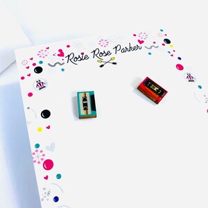 May include: A pair of cassette tape earrings. The earrings are made of wood and have a colorful design. The earrings are on a white card with the text "Rosie Rose Parker" written on it.