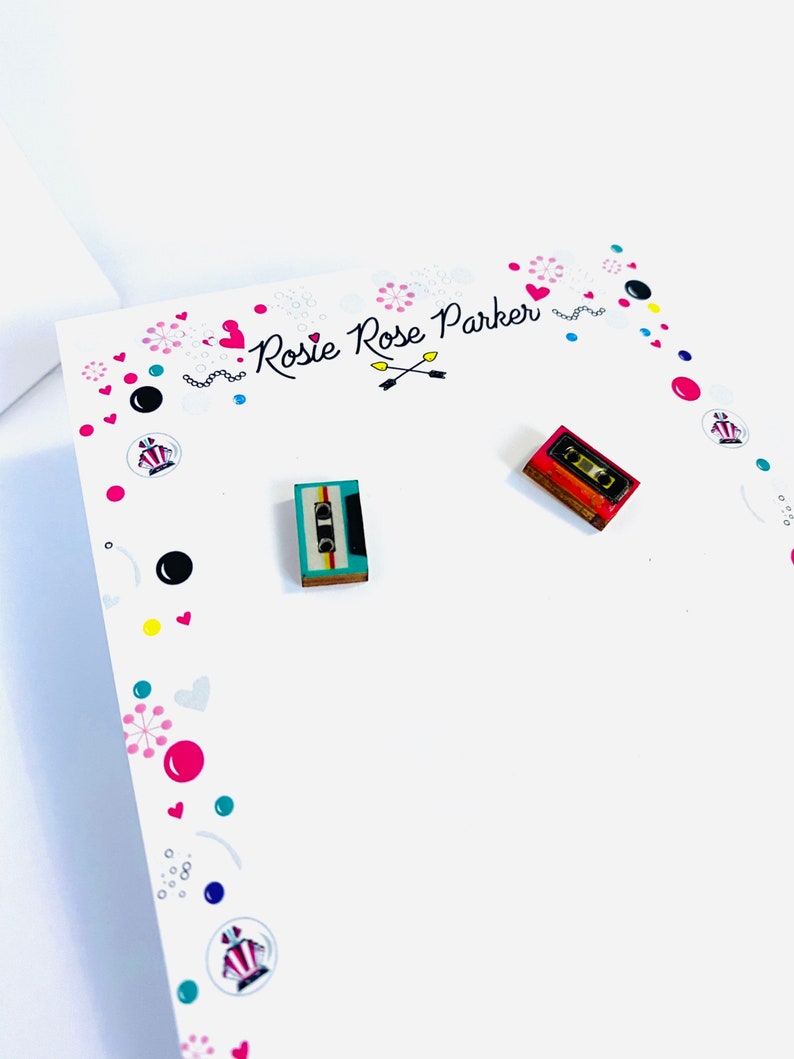 May include: A pair of cassette tape earrings. The earrings are made of wood and have a colorful design. The earrings are on a white card with the text "Rosie Rose Parker" written on it.