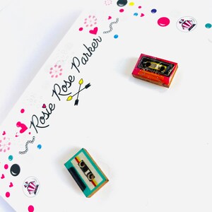 May include: A pair of small, wooden earrings shaped like cassette tapes. One earring is red with a yellow and white cassette tape, and the other earring is teal with a yellow and white cassette tape.