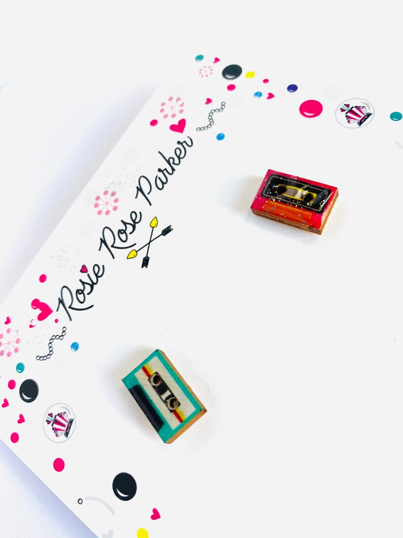 May include: A pair of small, wooden earrings shaped like cassette tapes. One earring is red with a yellow and white cassette tape, and the other earring is teal with a yellow and white cassette tape.