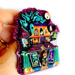May include: A glittery purple and black Halloween-themed house brooch with a spooky tree, ghosts, and pumpkins. The house has a door and windows with various Halloween decorations.