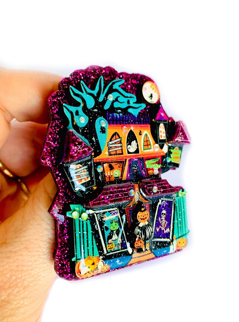 May include: A glittery purple and black Halloween-themed house brooch with a spooky tree, ghosts, and pumpkins. The house has a door and windows with various Halloween decorations.