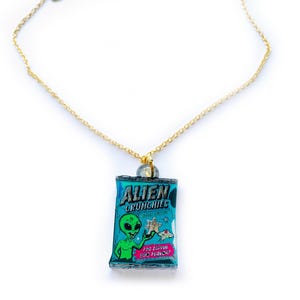 Könnte beinhalten: Eine goldfarbene Halskette mit einem Anhänger in Form einer Snacktüte. Die Tüte ist blau und zeigt eine Alien-Grafik mit dem Text "Alien Crunchies" und "Zoc Zappin BBQ Flavor!"