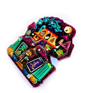 May include: A pink glitter Halloween-themed house pin with a spooky design. The house has a green roof, orange windows, and a black door. There are various Halloween decorations on the house, including pumpkins, bats, and ghosts.