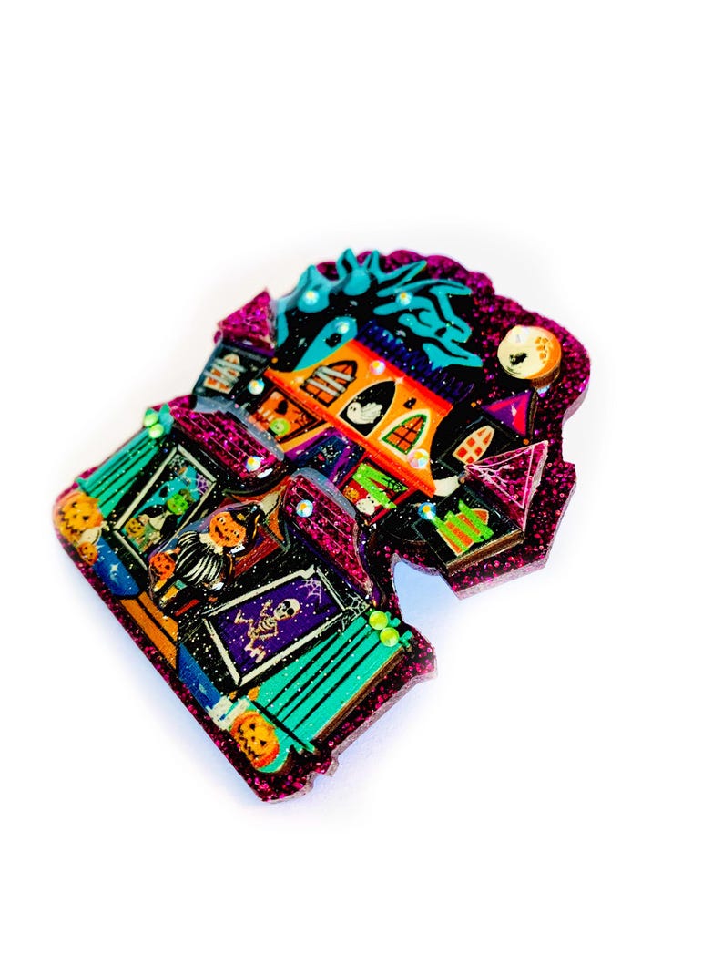 May include: A pink glitter Halloween-themed house pin with a spooky design. The house has a green roof, orange windows, and a black door. There are various Halloween decorations on the house, including pumpkins, bats, and ghosts.