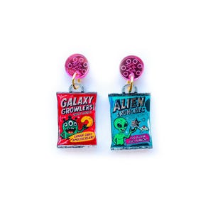 May include: A pair of novelty earrings featuring miniature chip bag designs. One earring is red with a green monster and the text "GALAXY GROWLERS". The other is blue with a green alien and the text "ALIEN CRUNCHIES". Each earring has a pink circular top.