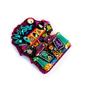 May include: A glittery pink and purple Halloween-themed house ornament with a black cat, ghosts, and pumpkins. The house is decorated with a variety of spooky details, including a spiderweb, a skull, and a cauldron.