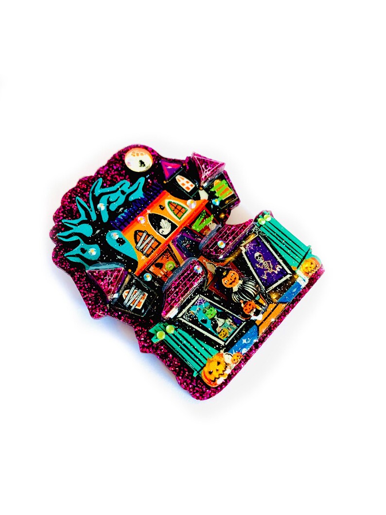 May include: A glittery pink and purple Halloween-themed house ornament with a black cat, ghosts, and pumpkins. The house is decorated with a variety of spooky details, including a spiderweb, a skull, and a cauldron.