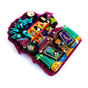 May include: A glittery, colorful Halloween-themed house brooch with a witch, ghosts, and pumpkins. The house is pink, green, and orange with black accents.