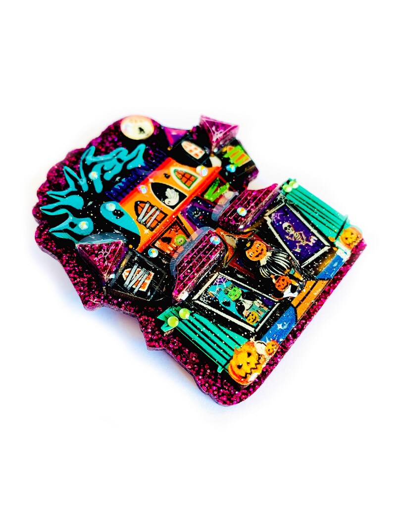 May include: A glittery, colorful Halloween-themed house brooch with a witch, ghosts, and pumpkins. The house is pink, green, and orange with black accents.
