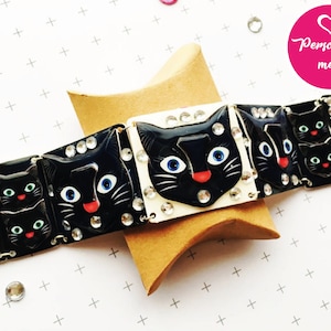 May include: A black and white bracelet with a repeating pattern of black cat faces with rhinestones. The bracelet is on a brown surface with a pink heart with the text "Personalise me!" in white.