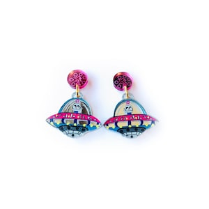Puede incluir: Un par de pendientes novedosos con un diseño de OVNI rosa y azul. Los pendientes tienen una parte superior circular rosa y una ilustración detallada de un platillo volante con una pequeña figura alienígena en el interior. Sobre un fondo blanco.