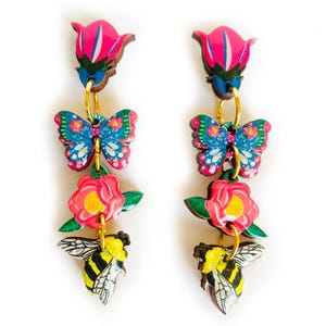 Könnte beinhalten: Ein Paar Hängeohrringe mit einer rosa Blume, einem blauen Schmetterling und einer gelben und schwarzen Biene. Die Ohrringe sind aus Holz und haben eine goldfarbene Oberfläche.