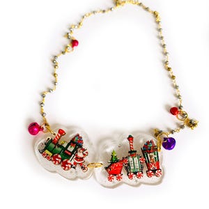 Può includere: Una collana con due ciondoli in resina trasparente e glitterata a forma di nuvola, ciascuno contenente l'illustrazione di un trenino natalizio con Babbo Natale. La catena è decorata con piccole perline metalliche e vari campanellini colorati.