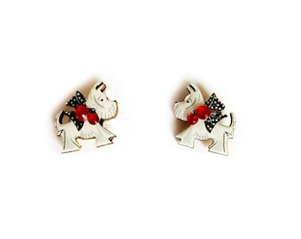 May include: A pair of white ceramic earrings shaped like a Scottish terrier dog with red and black accents.