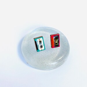 May include: A pair of small cassette tape earrings. One earring is red, black, and white. The other earring is teal, black, and white.