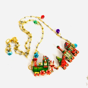 Può includere: Una collana con catena dorata e un ciondolo a forma di treno di Natale. Il treno è verde, rosso e bianco con una figura di Babbo Natale sul davanti. La catena ha perline colorate.