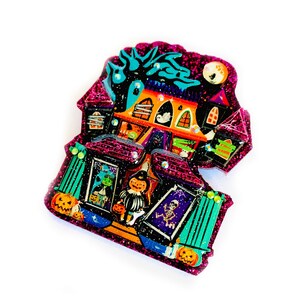 May include: A glittery pink and purple Halloween-themed house with a black cat, a ghost, and a skeleton. The house has orange and green windows and a black door. There are pumpkins and a witch in front of the house.