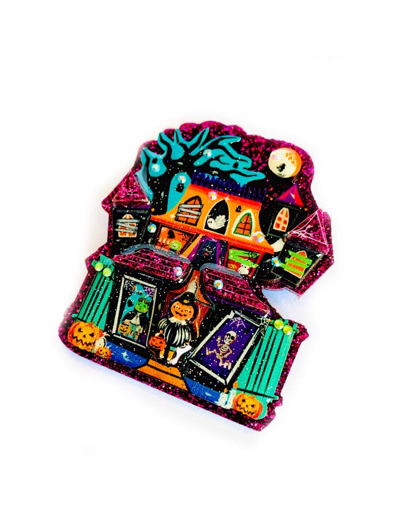 May include: A glittery pink and purple Halloween-themed house with a black cat, a ghost, and a skeleton. The house has orange and green windows and a black door. There are pumpkins and a witch in front of the house.