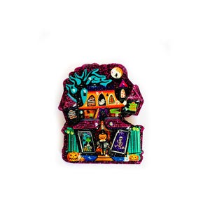 May include: A glittery purple and pink Halloween-themed house with a black cat, a ghost, and a witch. The house has a green roof and windows, and there are pumpkins and other Halloween decorations around it.
