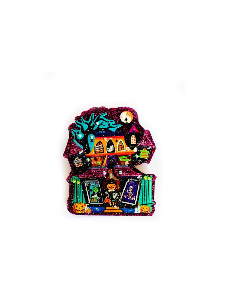 May include: A glittery purple and pink Halloween-themed house with a black cat, a ghost, and a witch. The house has a green roof and windows, and there are pumpkins and other Halloween decorations around it.
