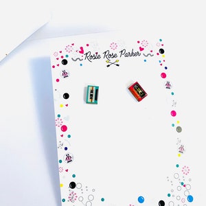 May include: A white card with a pink, black, and blue design. The card has the text "Rosie Rose Parker" and two small cassette tape earrings, one red and one blue.