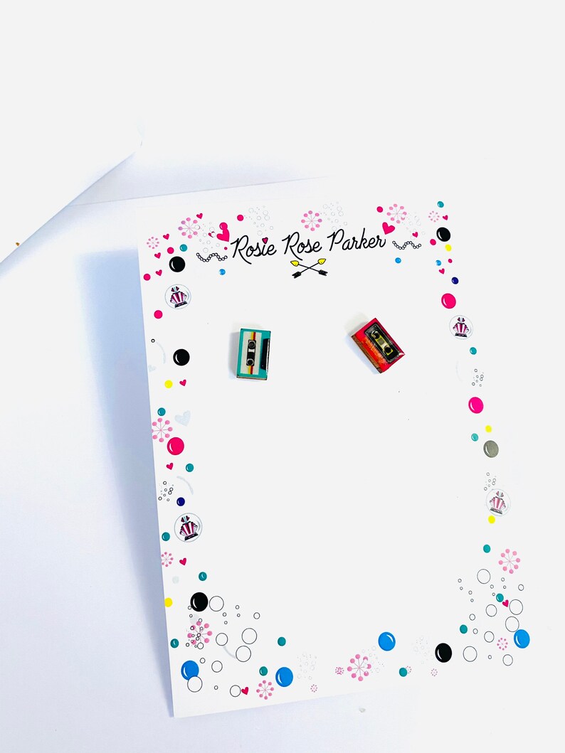 May include: A white card with a pink, black, and blue design. The card has the text "Rosie Rose Parker" and two small cassette tape earrings, one red and one blue.
