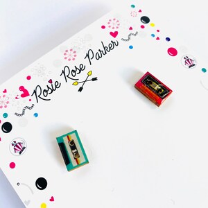 May include: A pair of cassette tape earrings. One earring is red, white, and blue. The other earring is turquoise, yellow, and black. The earrings are on a white card with the text "Rosie Rose Parker" in black.