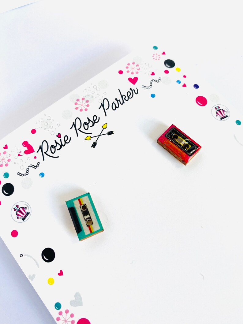 May include: A pair of cassette tape earrings. One earring is red, white, and blue. The other earring is turquoise, yellow, and black. The earrings are on a white card with the text "Rosie Rose Parker" in black.