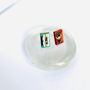 May include: A pair of cassette tape earrings, one red and one blue and green, on a white glitter background.