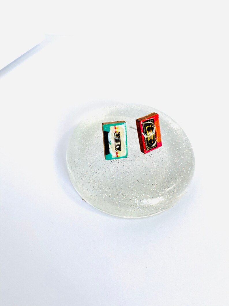 May include: A pair of cassette tape earrings, one red and one blue and green, on a white glitter background.