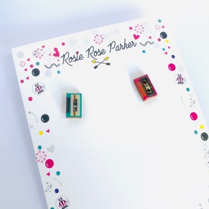 May include: A pair of small, colorful cassette tape earrings. The earrings are on a white card with the text "Rosie Rose Parker" and a heart design.