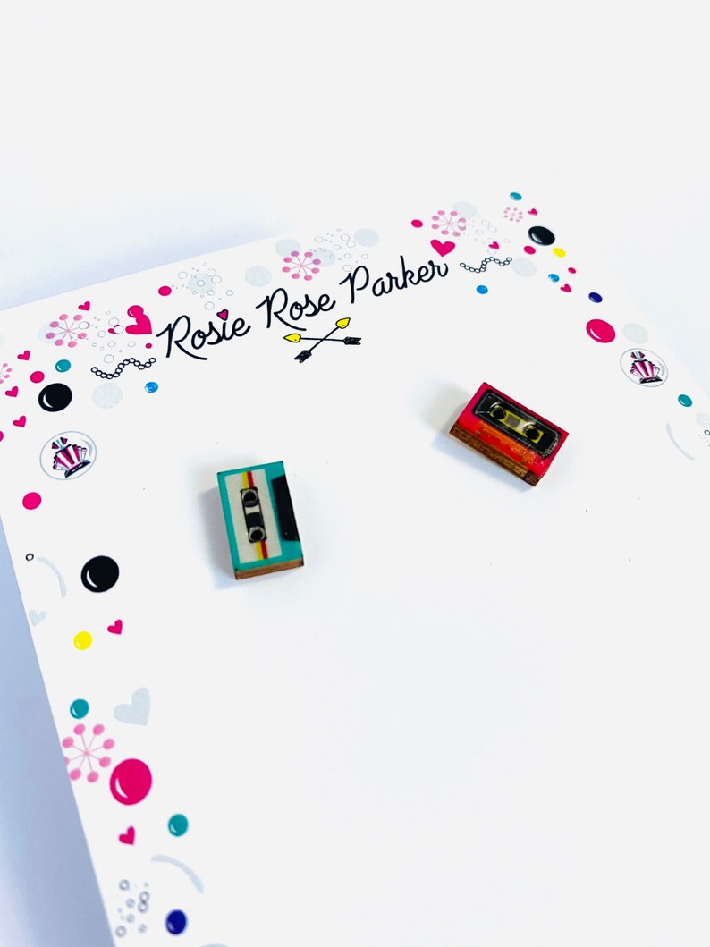 May include: A pair of wooden earrings shaped like vintage cassette tapes. One earring is teal with black and yellow details, and the other is red with black and white details. The earrings are displayed on a white card with a colorful design and the text "Rosie Rose Parker".