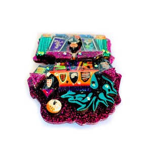 May include: A pink, green, and black glitter resin ornament with a colorful, detailed design. The ornament features a variety of shapes and patterns, including a castle, a dragon, and a crown.