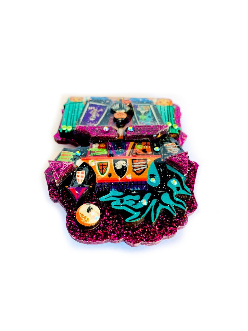 May include: A pink, green, and black glitter resin ornament with a colorful, detailed design. The ornament features a variety of shapes and patterns, including a castle, a dragon, and a crown.