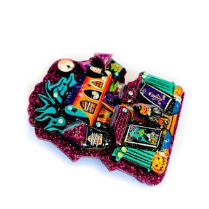 May include: A glittery pink and green Halloween-themed house brooch with a spooky design. The house has a black roof, orange walls, and a green door. There are ghosts, pumpkins, and other Halloween decorations on the house.