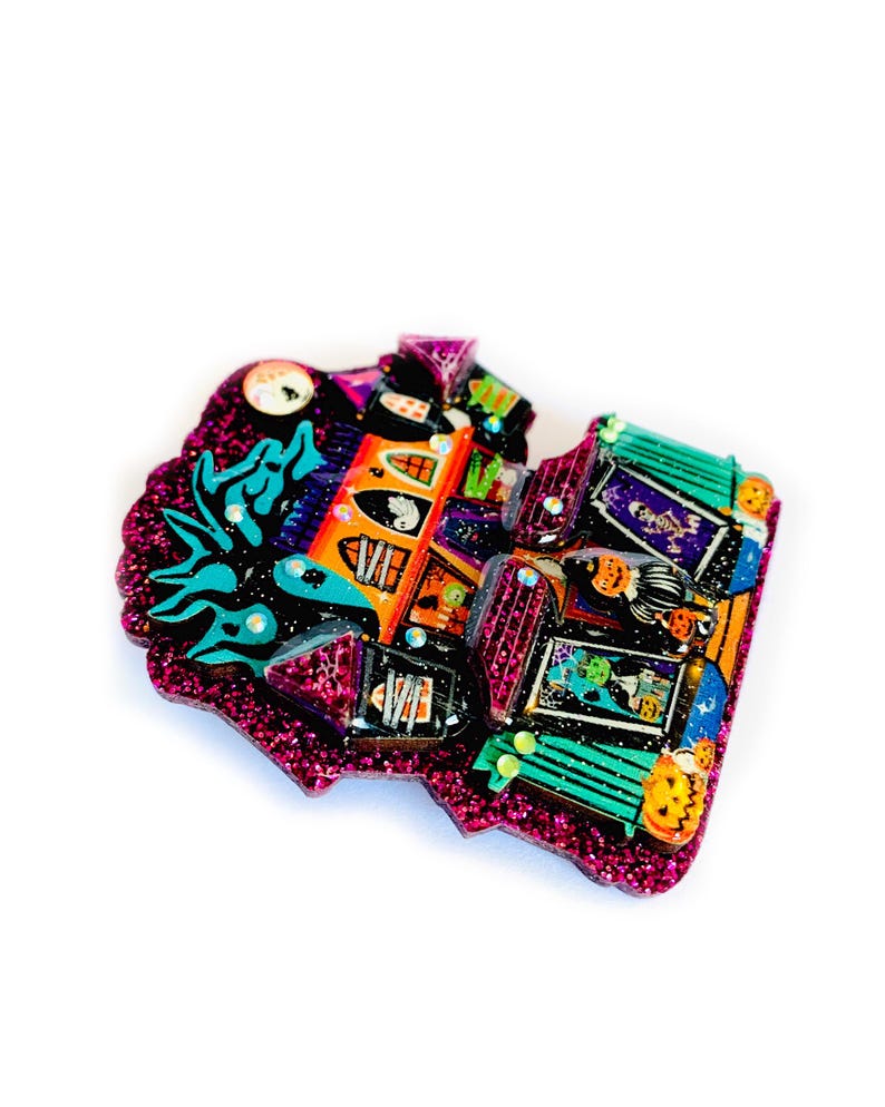May include: A glittery pink and green Halloween-themed house brooch with a spooky design. The house has a black roof, orange walls, and a green door. There are ghosts, pumpkins, and other Halloween decorations on the house.