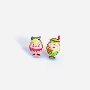 May include: A pair of wooden earrings shaped like cartoon eggs. The egg on the left is pink with a blue bow and the egg on the right is green with a yellow hat.