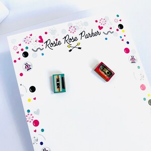 May include: A white card with the text "Rosie Rose Parker" and a pair of cassette tape earrings. One earring is red and the other is blue and green.