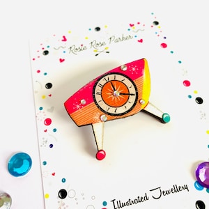 May include: A pink, orange, and yellow wooden clock brooch with rhinestones. The clock face is orange with black numbers and hands. The brooch is shaped like a retro clock with three legs. The brooch is on a white card with the text "Rosie Rose Parker" and "Illustrated Jewellery".