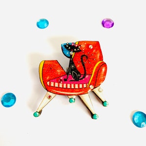 Puede incluir: Un gato negro sentado en una silla roja y blanca de estilo moderno de mediados de siglo con un patrón de estrella azul y blanco. La silla tiene tres patas con acentos turquesa.