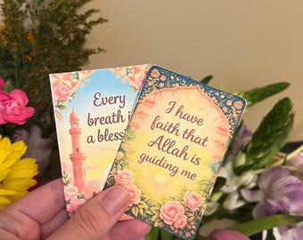 Può includere: Due biglietti decorativi con bordi floreali e testo. Uno dice "Every breath is a blessing" con un'illustrazione di una moschea. L'altro dice "I have faith that Allah is guiding me". I biglietti sono tenuti davanti a una composizione floreale.