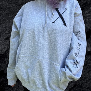 Pode incluir: Um moletom cinza claro com um grande "X" preto na frente e símbolos astrológicos na manga esquerda. O moletom tem capuz com cordão e um bolso frontal. O texto "My Life, My Hope" também está na manga.