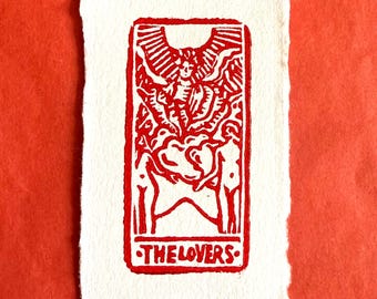 May include: A rectangular off-white paper print featuring a red ink block print illustration of The Lovers tarot card. The design depicts two figures beneath a winged angel. The text THE LOVERS is printed at the bottom, with APARADRAWS below it.