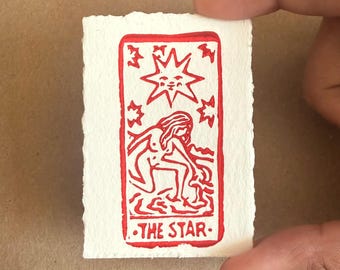 May include: A red and white tarot card print of "The Star" held by a hand. The card features a nude figure, a star, and the text "THE STAR". The card has a textured, handmade paper appearance.