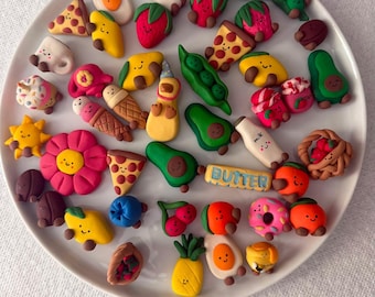 Può includere: Una collezione di piccoli ciondoli in argilla polimerica colorata a forma di alimenti, tra cui fette di pizza, avocado, fragole, piselli, cartoni del latte e frutta, tutti con semplici faccine sorridenti dipinte a mano su un piatto bianco.