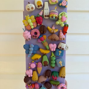 Può includere: Un'unità di archiviazione rettangolare color viola chiaro decorata con numerose piccole figurine in argilla colorate con volti sorridenti. Gli oggetti includono forme di cibo come una bottiglia di latte con l'etichetta 'milk', frutta, verdura, ciambelle, fiori e animali.
