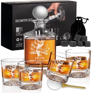 May include: A golf-themed decanter set with a golf ball stopper, a decanter with a golfer etching, and four rocks glasses. The set includes a black box and a bag of whiskey stones. The decanter reads "JECK BEST GOLFERS 202".