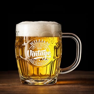 May include: A clear glass beer mug filled with golden beer and a thick white foam head. The mug has a handle and a vintage-style design that reads "William, Limited Edition 1974, Vintage, Aged to Perfection."